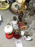 Seven miniature oil lamps arranged on a table surrounded by other dishware and items. Shows different shapes, colors, styles including red, white, amber, and decorative ones.