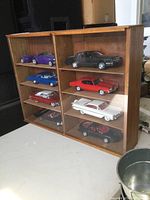 Front view of the wooden display case with glass sliding doors showing all 8 model cars inside across 4 shelves.