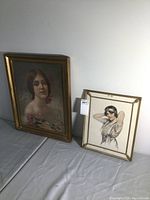Two framed prints side by side on a flat surface showing women portraits, one larger with a gold frame and the other smaller in a metal frame.