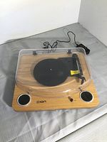 Top and angled view of Ion turntable with dust cover closed, showing wooden finish, built-in speakers, and power adapter.