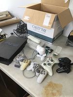 PS2 console, several controllers and cables on table, including a white PS One console in the background and a cardboard box.