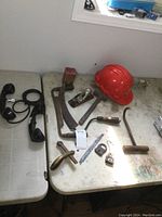 Wide view of various vintage tools including draw tool, sickle, plane, hammer, locks, hook tool, phone handsets, and hard hat on table.
