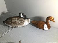 Two carved wood waterfowl decoys placed on a light surface, one painted with weathered white and gray paint representing a goose, the other in natural wood finish representing a duck.