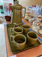 Photo showing green ceramic coffee/tea set with pot on warmer stand, four cups and tray arranged on a table with other kitchen items in background.