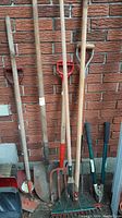 Photo showing long-handled shovels, garden forks, fan rake, hoe, standard rake, and heavy clippers leaning against a brick wall.
