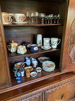 Wood cabinet shelf with assorted glassware, porcelain tea cups, plates, and ceramic jugs.