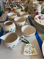 Photo of ceramic mugs, small vase, creamer, sugar bowl, and bell-shaped ornament all decorated with shamrocks and gold trim, arranged on a surface with blue tape.