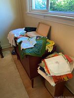 Lot of assorted linens arranged on wooden furniture by window, including placemats and napkins.