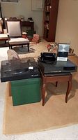 Telefunken record player, Mitsubishi VCR, RCA cassette player on small table and green ottoman in basement room with upholstered chairs in background.