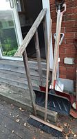 Photo shows three shovels with wooden handles leaning against a wooden railing beside a house door. One shovel has a red metal scoop blade, another shovel blade is silver and metal, the third shovel has a large black plastic snow shovel blade. A wooden push broom is also visible.