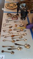 Photo showing twenty different souvenir spoons arranged in rows, two tea strainers placed near the spoons, small dark metal bud vase, and small pitcher with handle and hinged lid on a flat surface.