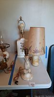 Three table lamps on white table; porcelain base lamp with floral design, stone base lamp with parchment shade, and stone base lamp without shade.