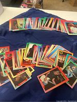 Wide view showing a large assortment of colorful collectible cards laid out across a blue cloth. The cards feature different hair band rock stars and bands with vibrant borders in various colors.