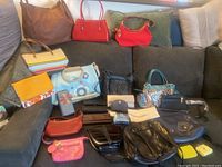 Full view of the assortment of handbags and wallets arranged on a dark sectional couch showing variety of colors, patterns, and materials.
