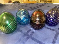 Four colored Hungarian crystal eggs in green, light blue, dark red, and dark blue with distinct etched patterns displayed on blue fabric.