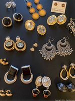 Overview of 12 pairs of mixed vintage clip-on and modern pierced earrings with various shapes, materials, and colors including gold-tone, silver-tone, black resin, and crystal rhinestones.