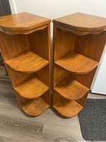 Front perspective of two solid wood corner shelves showing three tiers each with rounded front edges and a flat top surface. Shelves finished in medium brown wood tone.