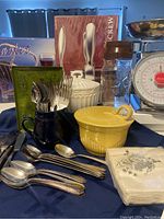Wide view of all kitchen items including boxed corkscrew and wine basket, lidded bowls, silver utensils, jars, creamer, and napkins