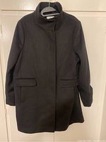 Full front view of the black wool blend coat hanging on a hanger showing the stand collar and flap pockets.