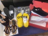 Lot of five pairs of women's shoes displayed on flat surface including yellow Crocs, black Lane 8 sneakers, black Shushop heels, black A.N.A thong sandals, and black Ulogu beaded sandals.