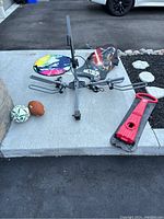 Photo showing metal hitch bike rack with attached clamps, skateboard, colorful circular disc, soccer ball, football, and red sled pull handle on concrete.