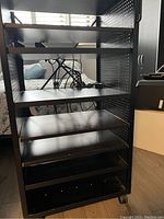 Front view of black metal utility cart showing six adjustable shelves and pegboard style side panels. Shelves are smooth and firmly mounted. Cart is positioned on wooden floor in room with window light behind.