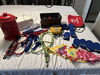 Photo showing assorted dog leashes in red, blue, and green, blue winter booties, colorful dog scarves, grooming tools, a yellow waste bag holder and a small black container with dog toys and grooming accessories all arranged on a white surface.