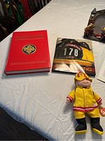 Red firefighter book, firefighter training book, yellow firefighter beanie doll on white table