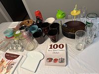 Wide view showing assortment of kitchen and drinkware items including glasses, mugs, containers, plates, teapot, and books on a white surface.
