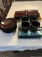 Set of 24 pieces including round and square plates in brown and blue, and dark brown mugs on a white tablecloth.