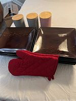 Eight dark brown ceramic square plates stacked, three pastel ceramic canisters with bamboo lids, and red oven mitts laid out on a white surface.