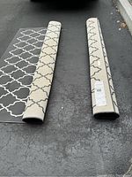 Two large rolled IKEA rugs with beige and white geometric trellis pattern partially unrolled on asphalt showing front and back sides.