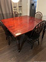 Square solid wood dining table with glossy reddish-brown top and four solid wood spindle-back chairs with dark stained legs around it, placed on wooden floor.