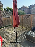 Photo of closed large red outdoor deck umbrella on a wooden deck, showing full length and base.
