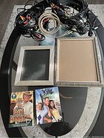 Overview photo showing a collection of tangled audio/video cables, two picture frames of different sizes and styles, and two DVD cases placed on a black glass table.