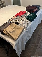 Photo showing various folded women's clothing items including pants, tops, and dresses in different colors and materials on a table.