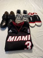 Photo of folded black Miami Heat basketball jersey with white and red lettering, a pair of black dress shoes with velcro straps, gray athletic shoes, and black, red, and white knit wristbands with 'Canada' text.