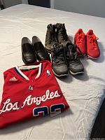 Four pairs of youth shoes and a folded red basketball jersey laid out on a bed, showing front of jersey with 'Los Angeles' text.