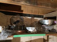 Baking pans, cake molds, cookware including metal Bundt cake pans and GreenPan pot, some wrapped in plastic, stored on basement shelf.