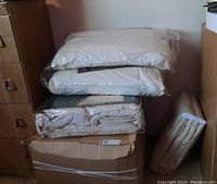 Stacked packaged bedding items: two pillows on top, folded duvet below, sealed box and rolled cover