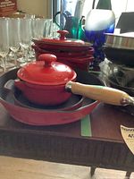 Photo showing multiple red enameled cast iron cookware pieces including two saucepans with lids and wooden handles, a skillet underneath, and a black trivet below the skillet.