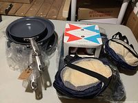 Set showing fry pans stacked with metal handles visible, adjacent boxed insulated glasses with watermelon design, and mesh storage bags.