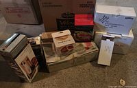 Lot of kitchen items in original boxes placed closely together on a concrete floor including Wolfgang Puck apple peeler, dough press set, Braun stick mixer box, and others.