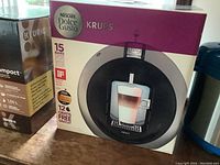 Front box image showing the Krups Nescafé Dolce Gusto coffee maker in black and grey with cup of layered coffee shown on front.