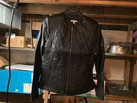 Black leather jacket hanging on a hanger in a basement storage area with visible embossed floral designs on the front.