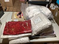Photo of assorted linens on a table including packaged tablecloth, white house coat, red floral fabric, and bedding items