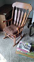 Full view of solid wood rocking chair with brown cushion on carpet next to stack of puzzle books.