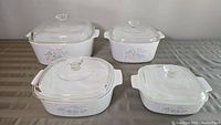 Four white Corningware casserole dishes with floral decorations and clear glass lids displayed on a table
