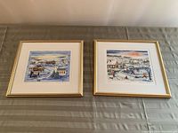 Two Christmas-themed art prints in gold frames on a beige surface showing snowy village scenes with people and buildings.