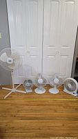 Photo showing the full lineup of five fans including the tall pedestal fan, medium desk fan, two smaller desk fans, and one small floor fan in front of white paneled doors on wooden floor.